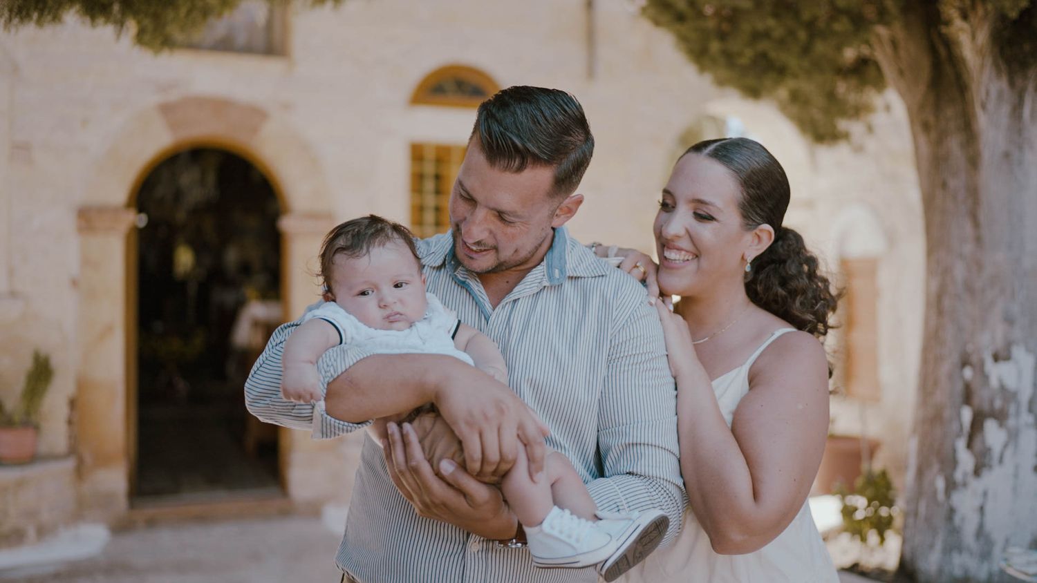 A loving family shares a tender moment together outside a rustic Mediterranean-style building on a sunny day.
