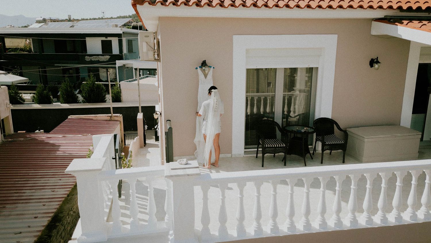 A person in a white dress stands on a Mediterranean-style terrace with white balustrades and rustic roof tiles.