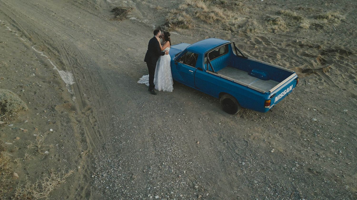 A couple stands next to a vintage blue pickup truck on a gravel mountain road high in the hills.