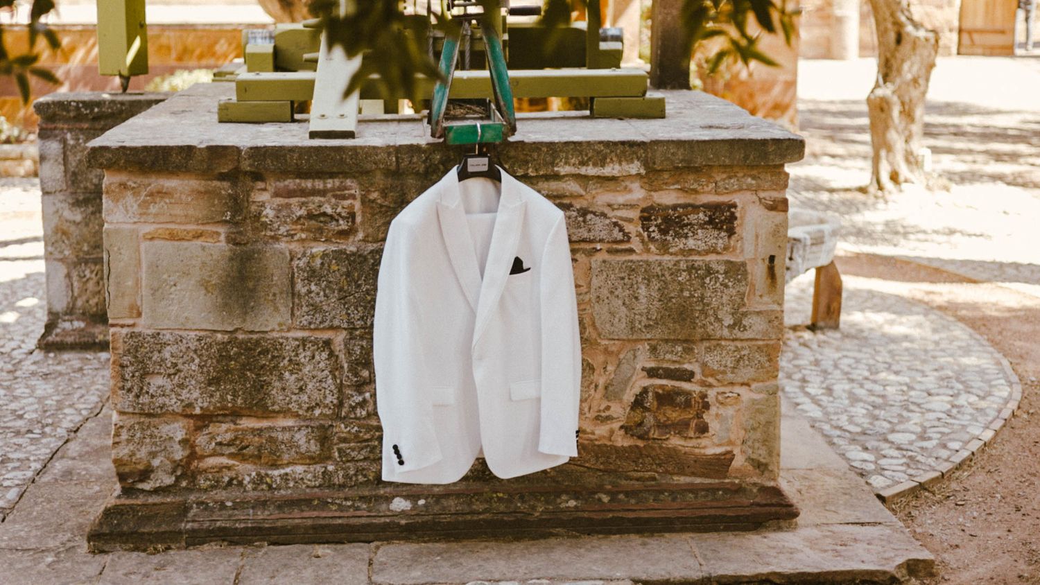 A white blazer hanging on a rustic stone fountain in a sunny Mediterranean courtyard setting.