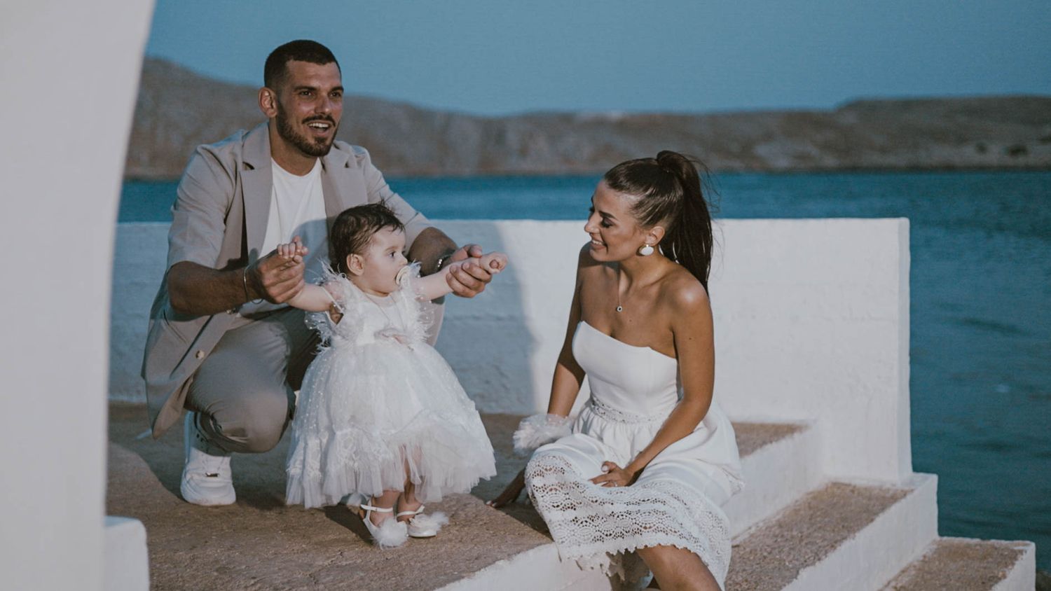 A family dresses in white sits together on seaside steps overlooking a scenic blue bay on a sunny day.