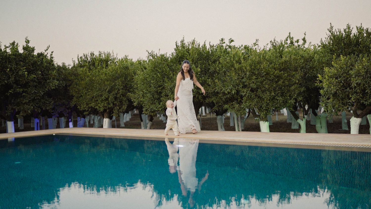 A bride in a white wedding dress stands at the edge of a luxurious swimming pool reflecting her image in the blue water.