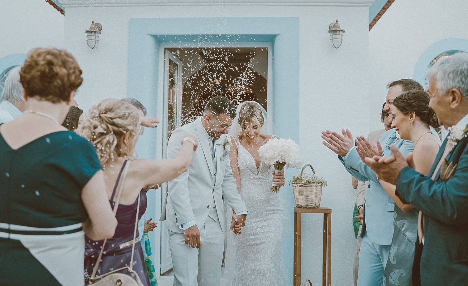 Wedding guests throw confetti on a newlywed couple outside a venue with a bright blue door.