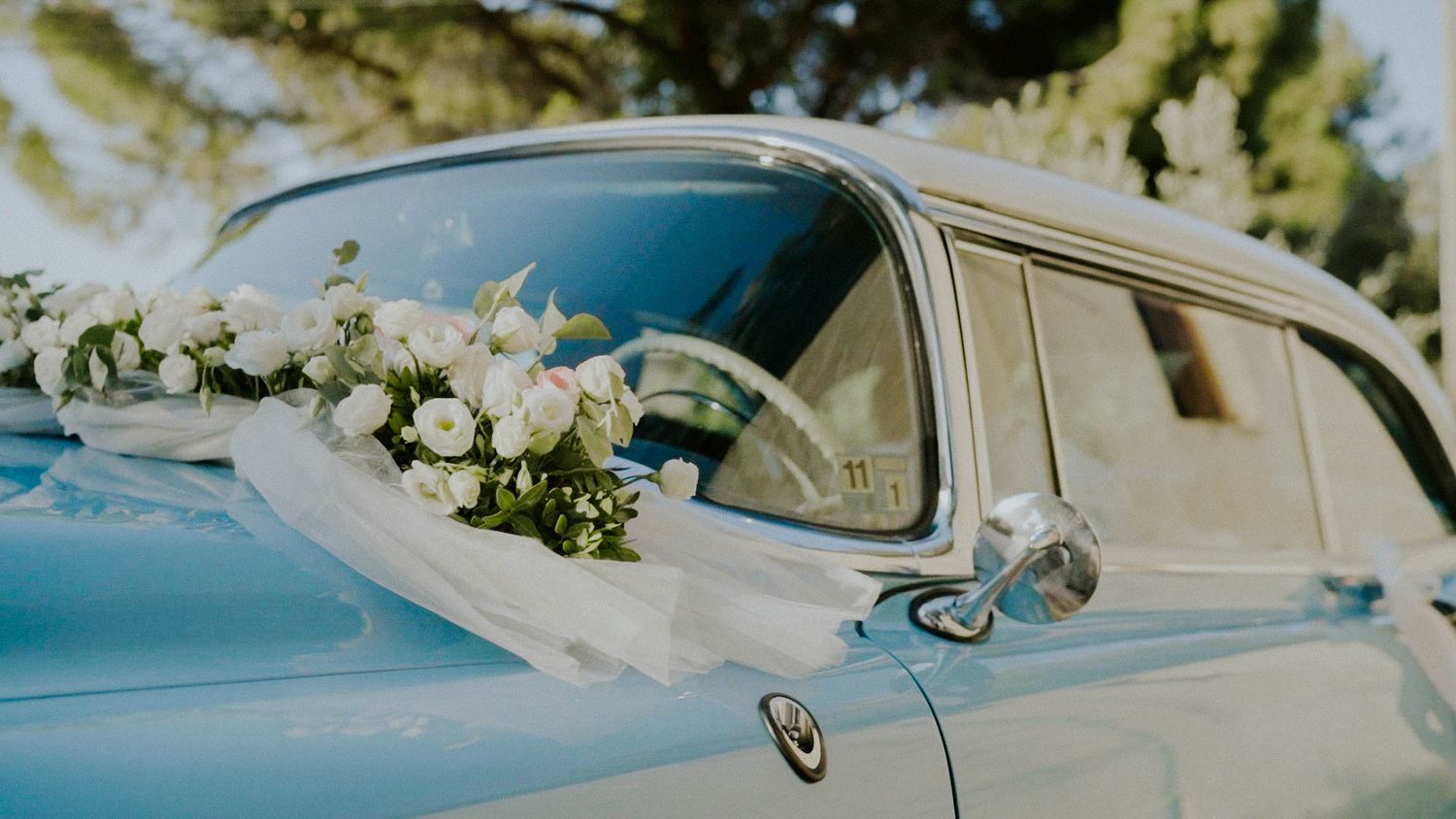 Wedding floral arrangements featuring white roses and greenery adorn a luxury vintage BMW convertible car at an outdoor ceremony.
