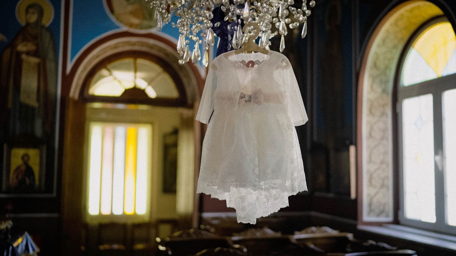 A delicate white lace baptism dress hangs from a chandelier in an ornate church interior with arched windows.