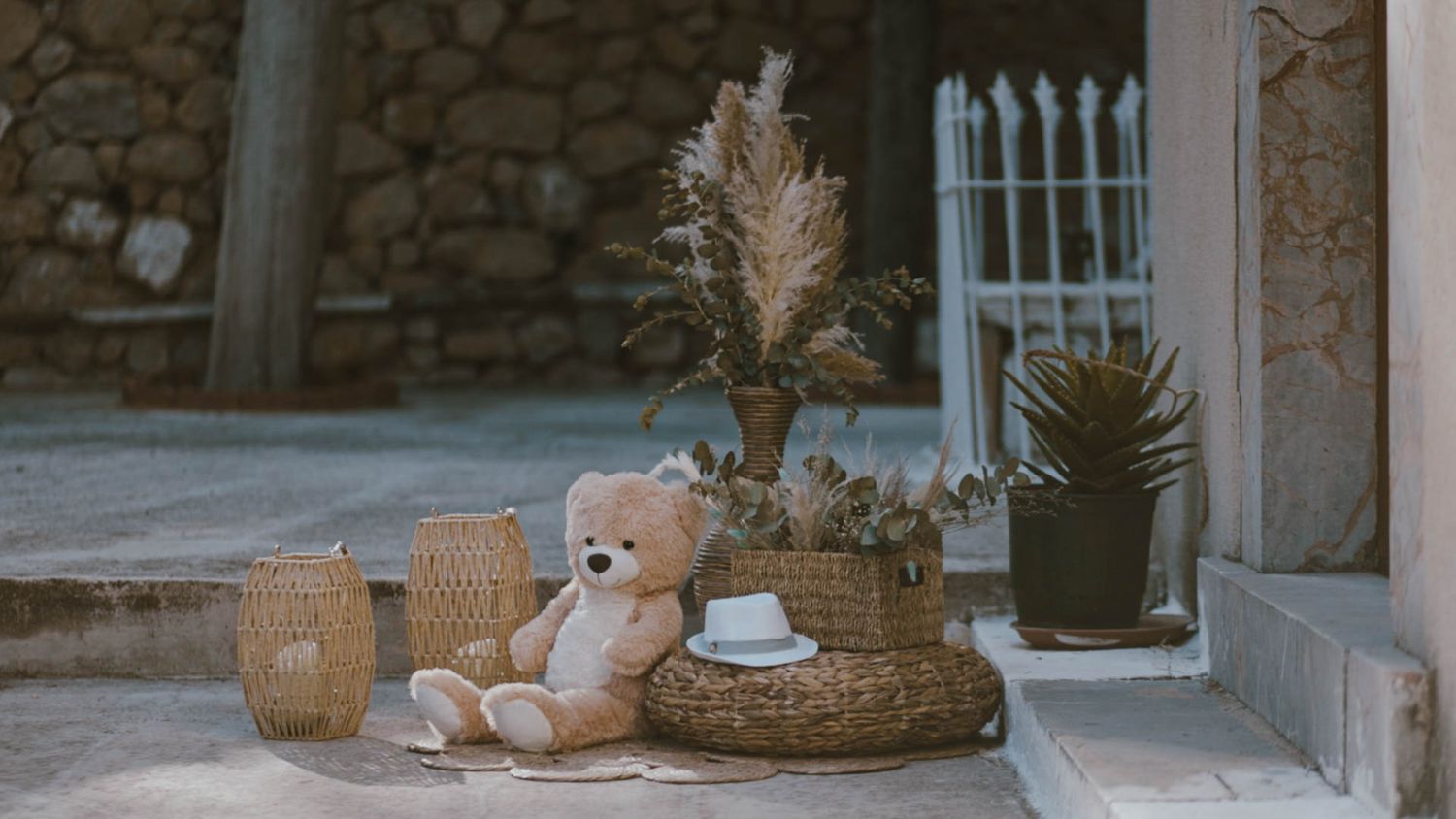A cozy outdoor patio scene with teddy bear, woven baskets, dried pampas grass, and potted plants against a stone wall.