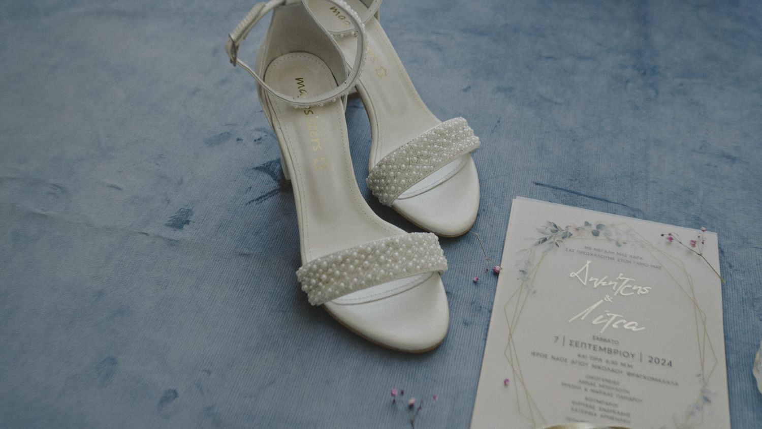 Elegant white satin wedding shoes with rhinestone embellishments displayed alongside formal invitation on blue surface.