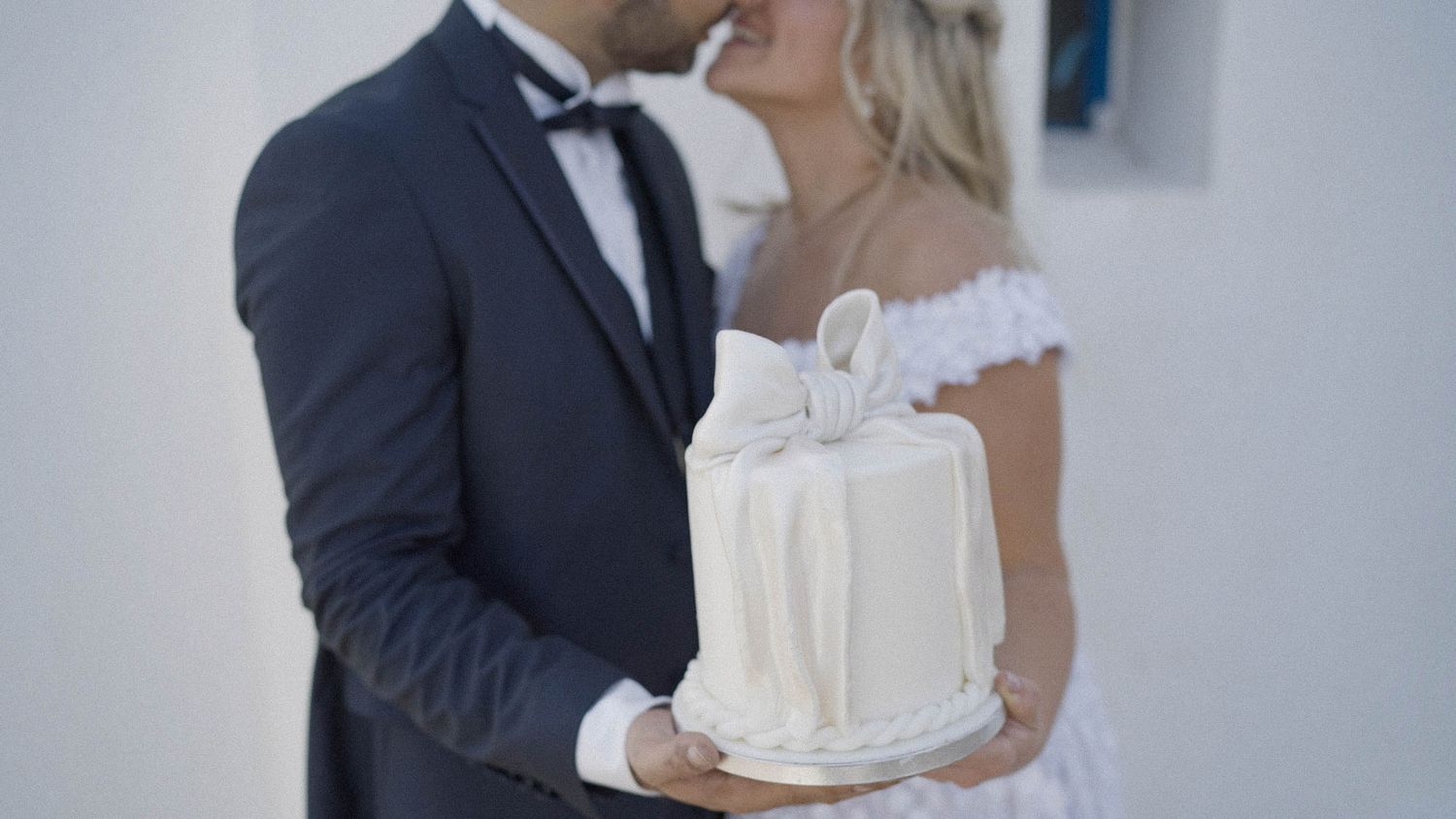 A bride and groom share an intimate moment while holding an elegant white wedding cake with pearl decorations.