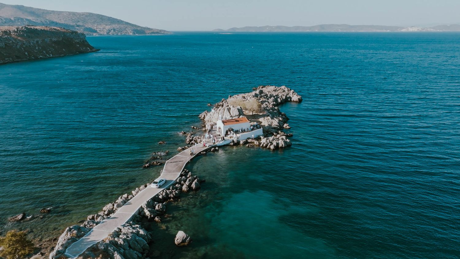 Aerial view of a curved stone bridge connecting to a small rocky island in turquoise Mediterranean waters.