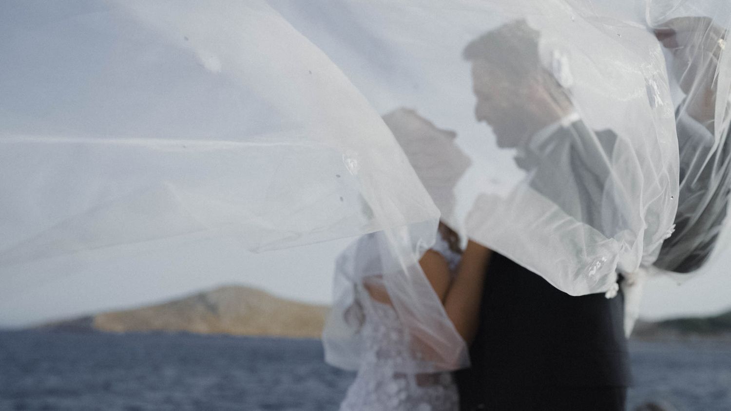 A flowing white veil blows in the wind against an ocean backdrop with mountains visible in the distance.