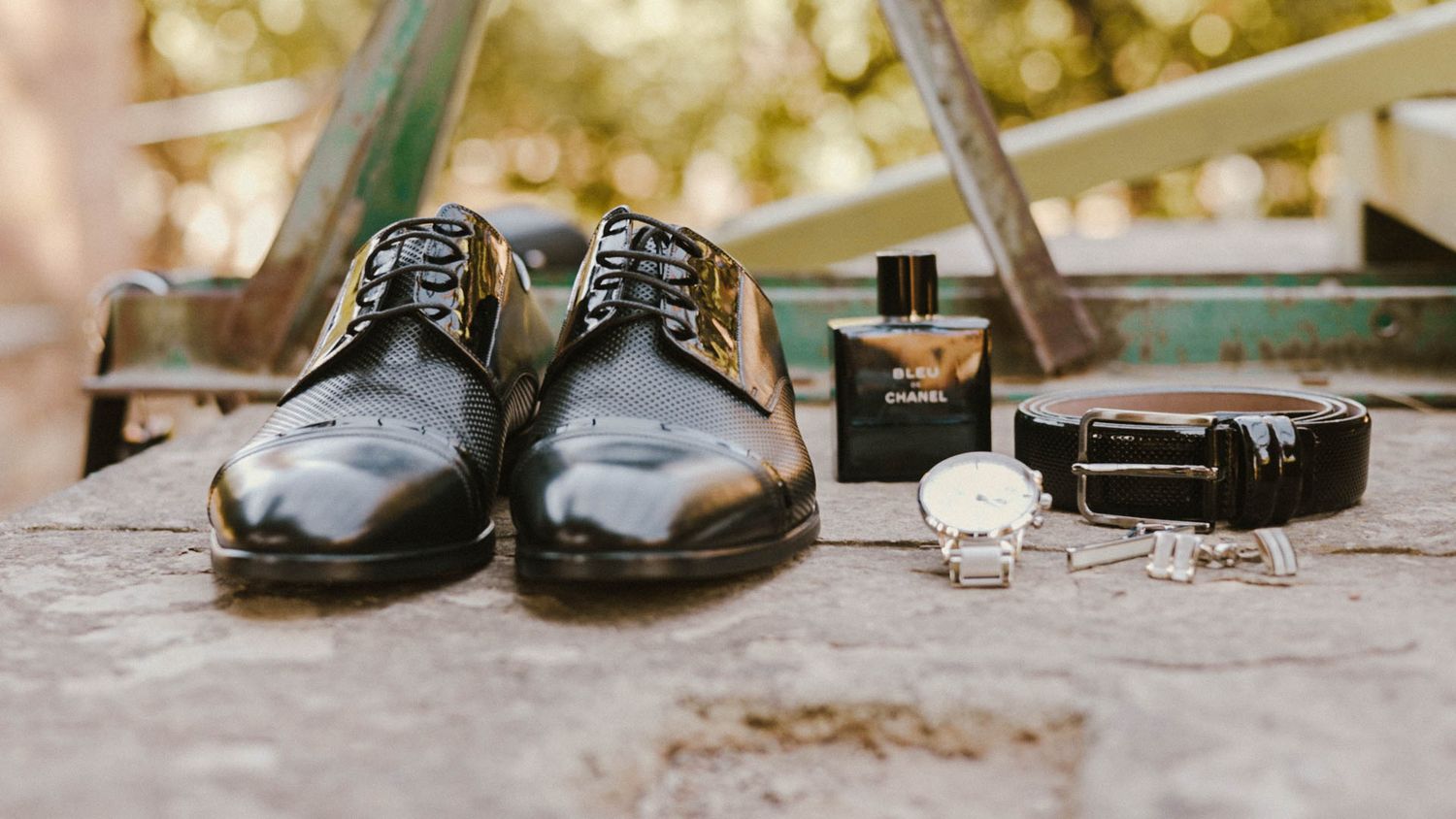 Shiny black dress shoes displayed with watch, cologne, cufflinks and belt on textured wood surface with bokeh background.