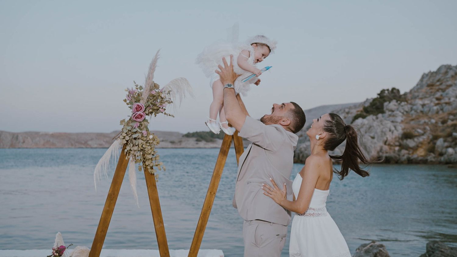 A couple celebrates by the water at sunset with a wooden arch decorated with flowers during their outdoor ceremony.