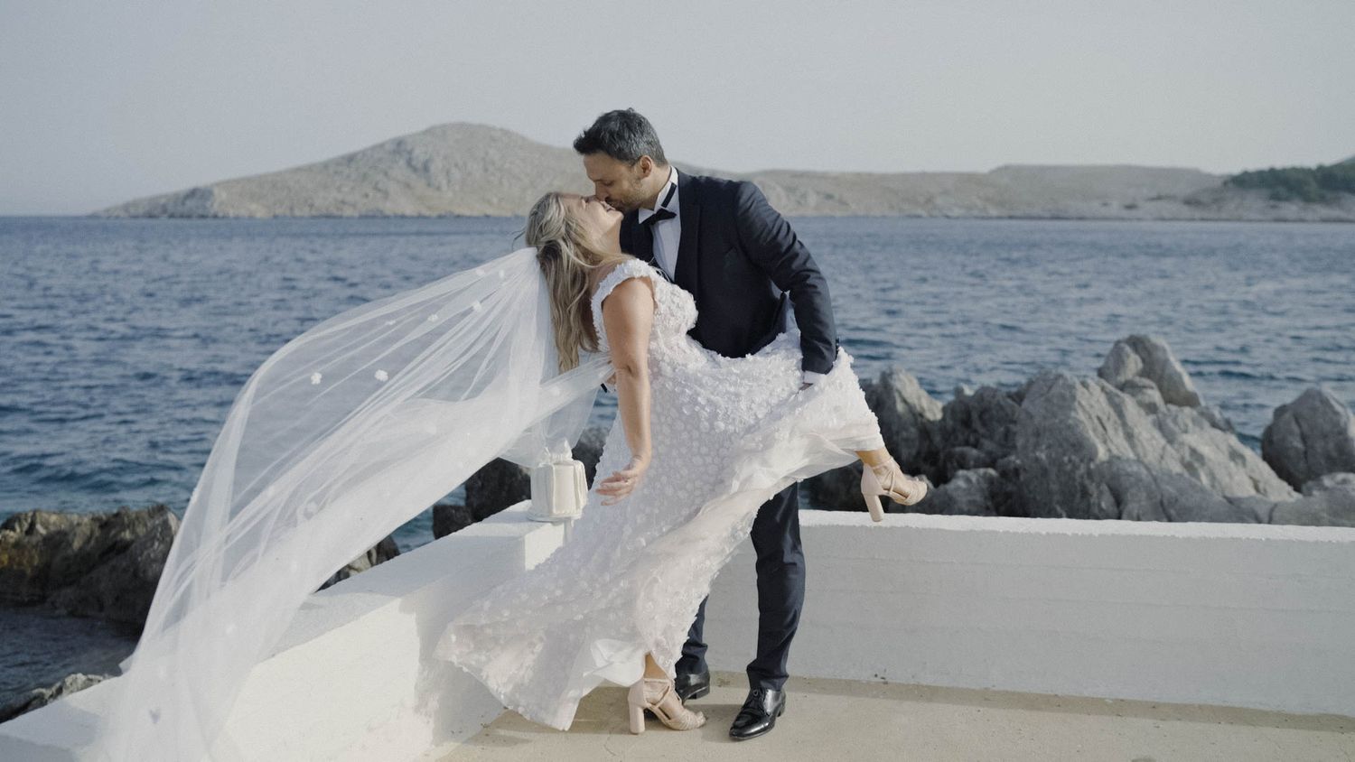 A romantic wedding couple embraces on a boat with a flowing white veil and rocky coastline in the background.