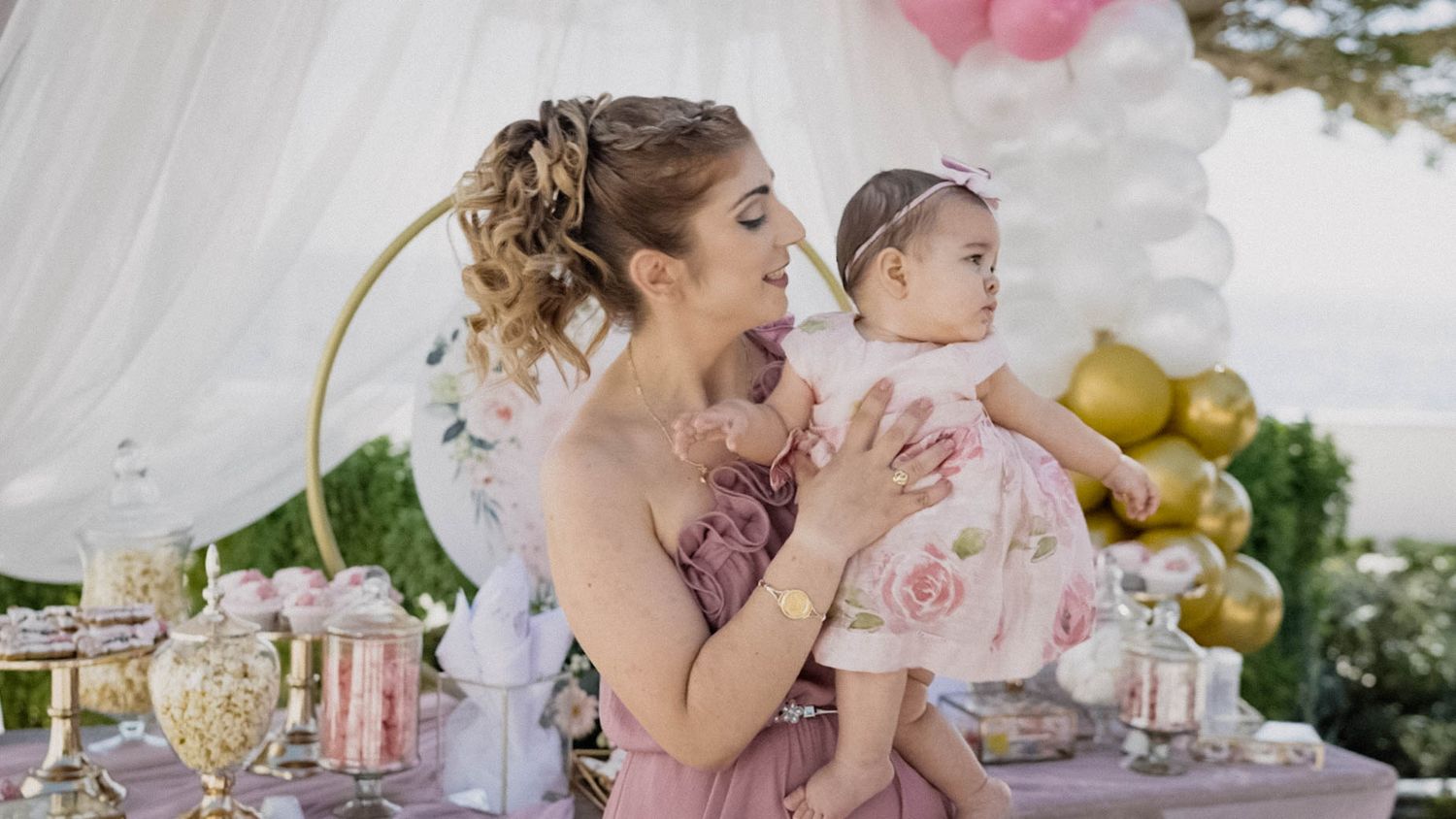 A tender moment at an elegant pink-themed outdoor party with decorative place settings and balloons in the background.