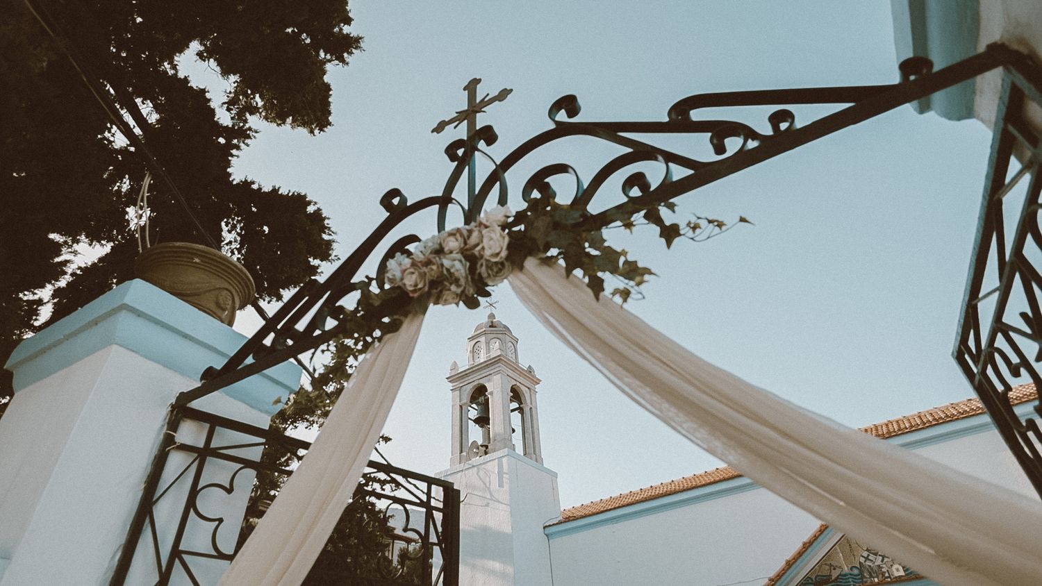 Elegant wedding arch with white fabric draping and ornate iron details frames a church steeple against a blue sky.