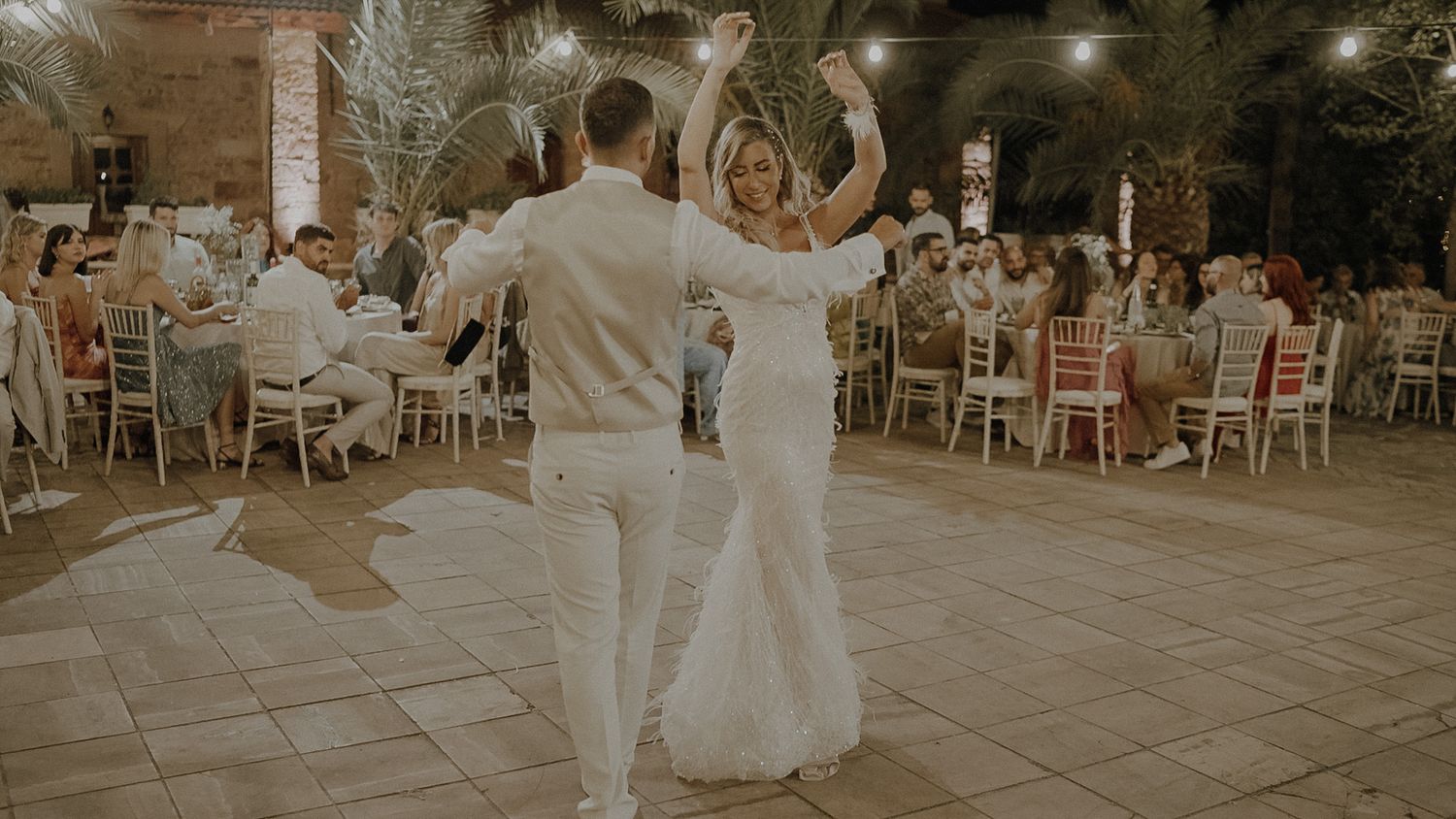 Two people in white attire dance together on a tiled outdoor patio at an evening wedding reception with tables in background.