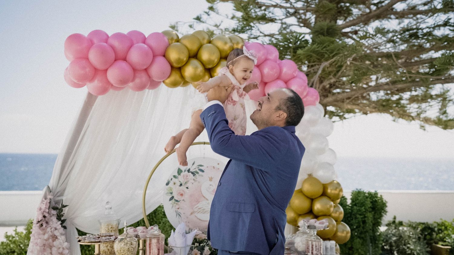 A couple shares a romantic moment under a pink and gold balloon arch at an outdoor wedding ceremony overlooking the ocean.