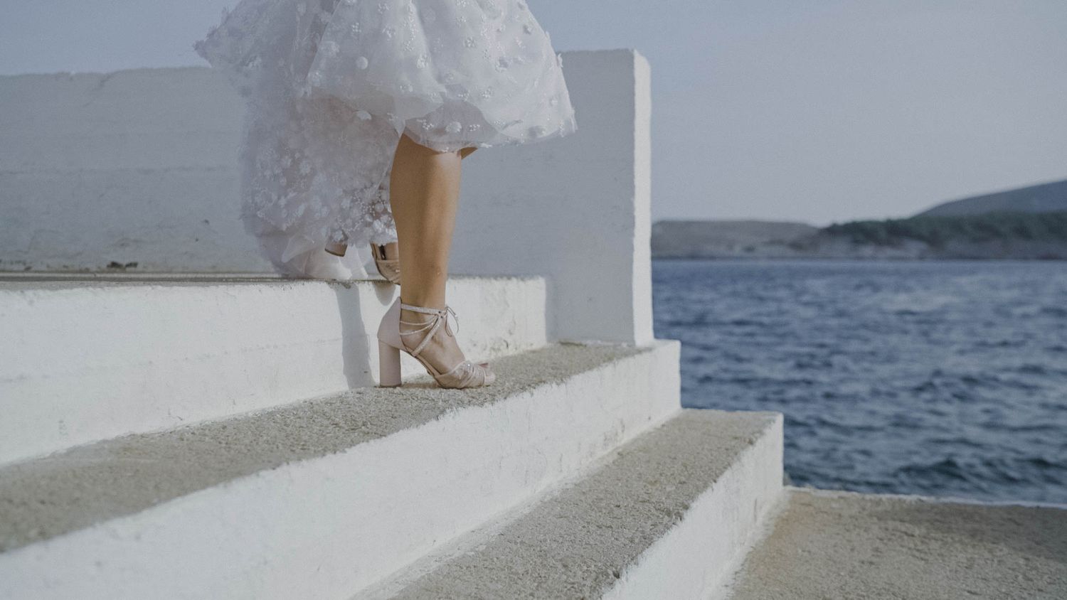 A person wearing a white lace dress and strappy heels stands on white stone steps overlooking a Mediterranean seascape.