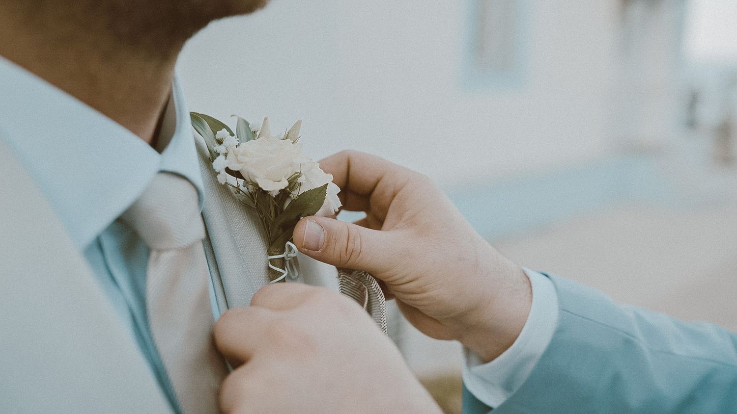 Someone in a light blue suit attaches a white boutonniere flower to their lapel during a formal event.
