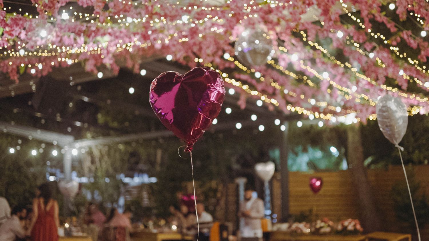 Pink heart balloon floats among twinkling string lights at an outdoor evening party setting.