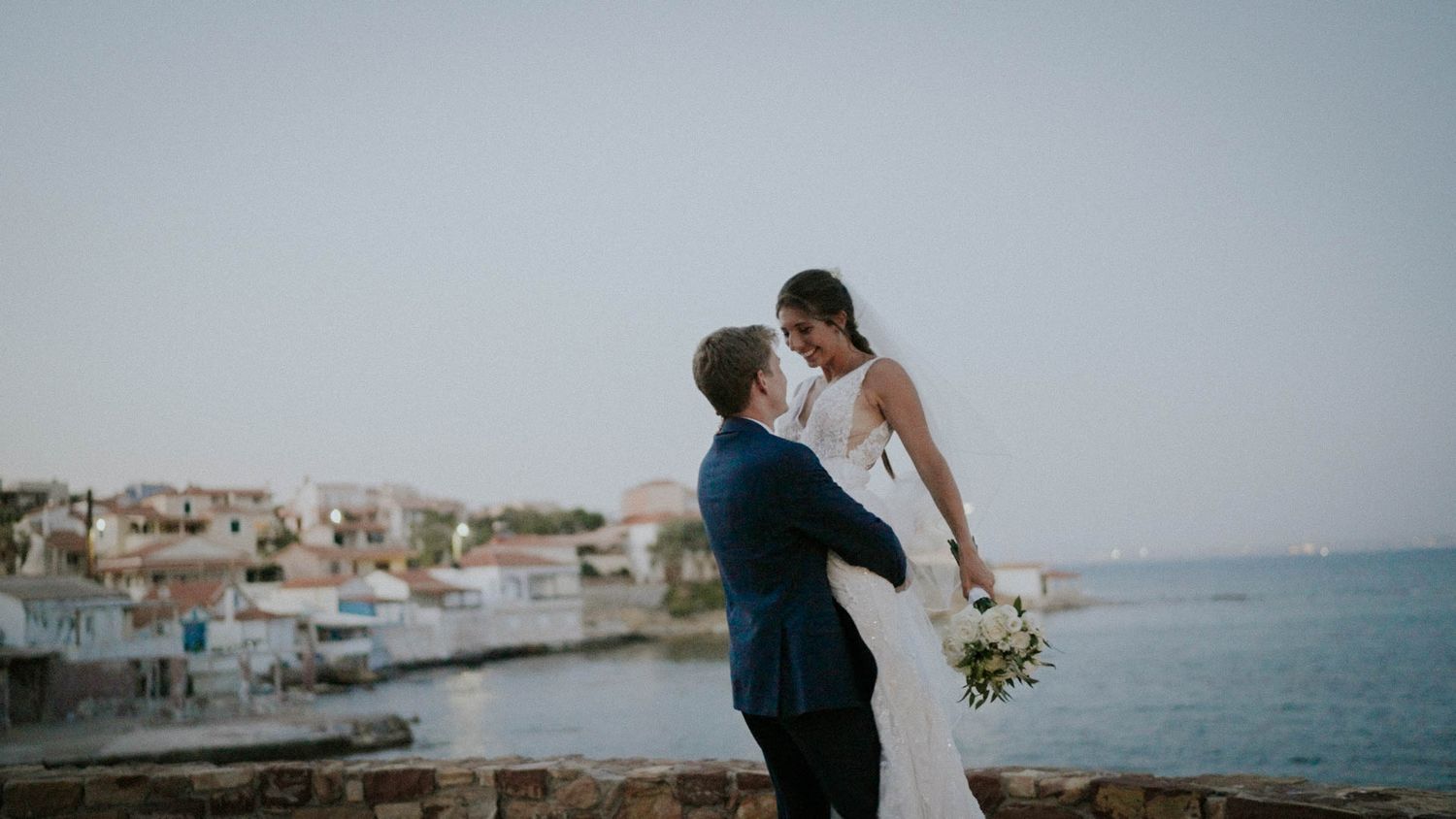 A romantic wedding couple embraces on a coastal overlook with a Mediterranean village and blue sea in the background.