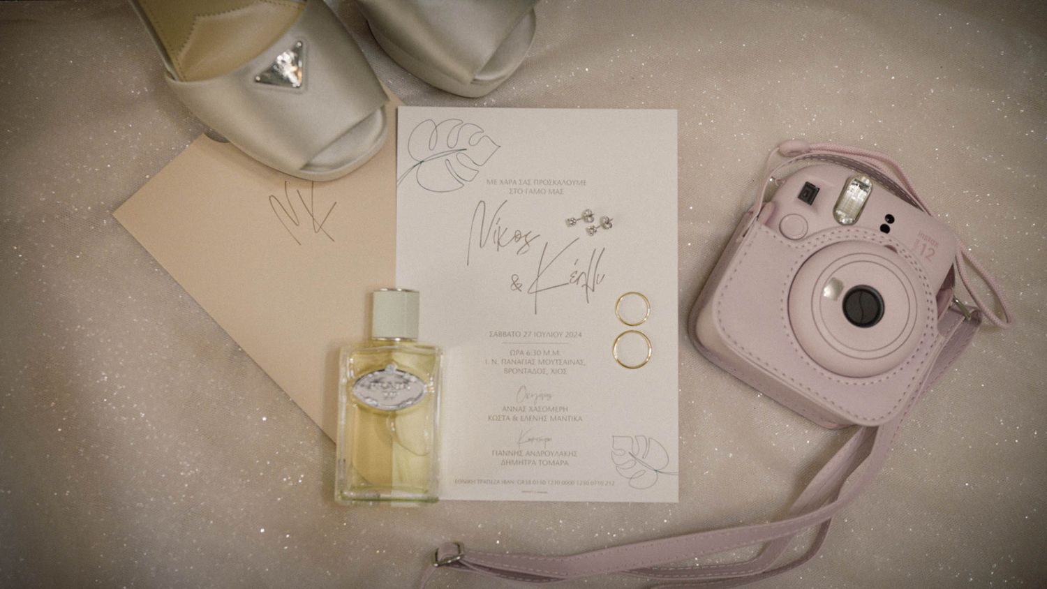 A wedding invitation lies beside a pink Instax camera and perfume bottle on a cream-colored surface.