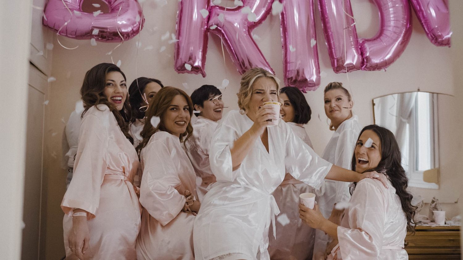 A group celebrates in matching white robes at a bridal preparation party with pink balloon decorations.
