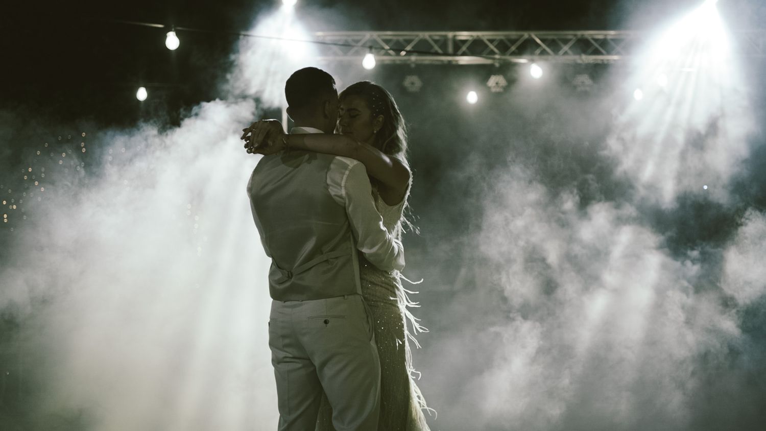 A couple embraces on a dark stage surrounded by dramatic fog and bright spotlights during a performance.