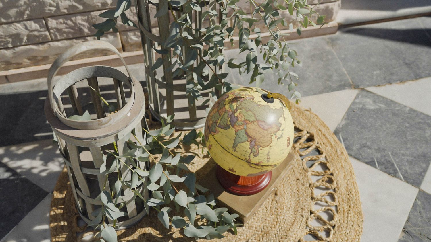 Vintage-style glass globe and lantern displayed with eucalyptus on stone surface in sunlit outdoor setting.