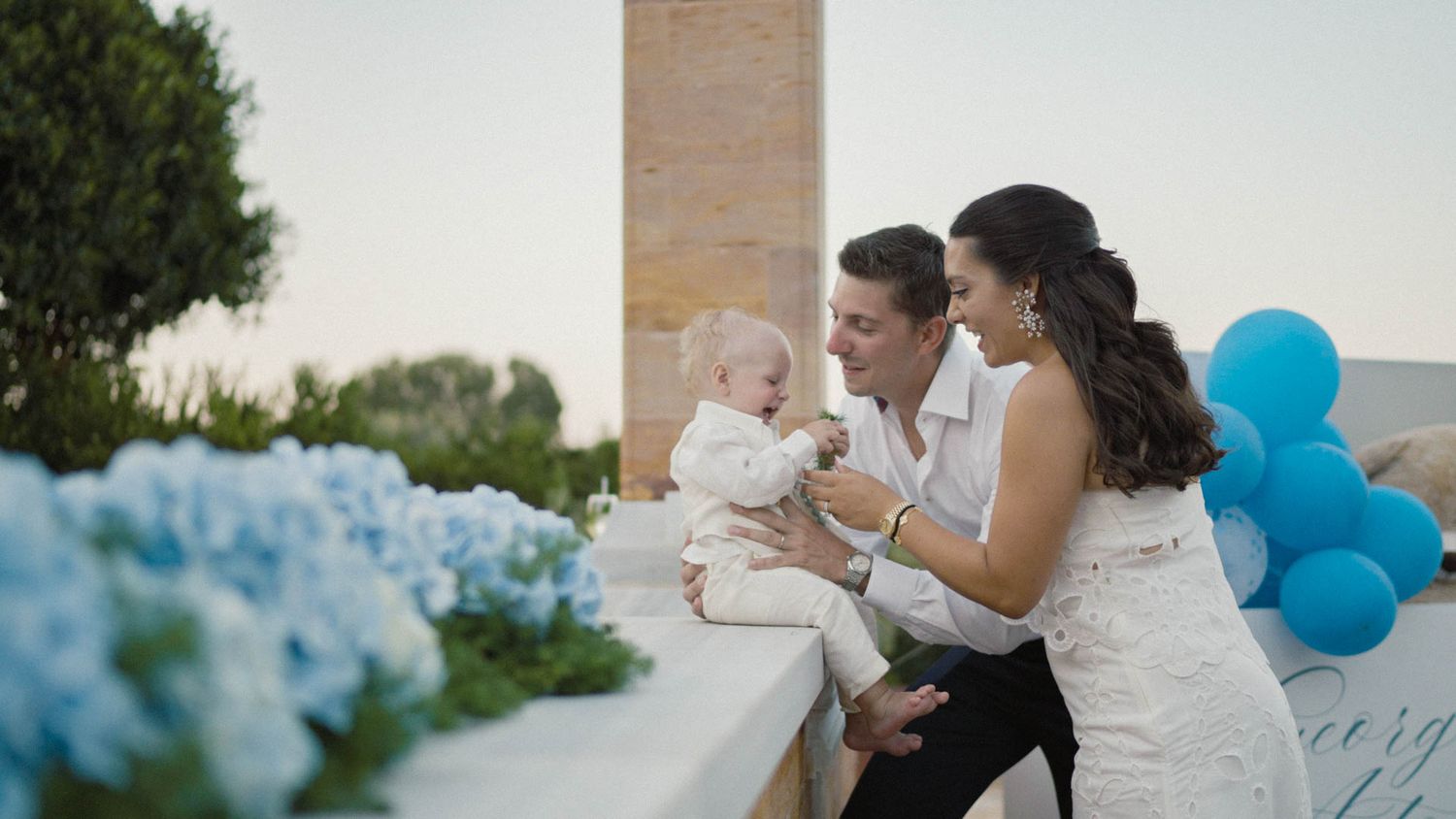 A couple in wedding attire sit at an outdoor venue decorated with blue hydrangeas and balloons.