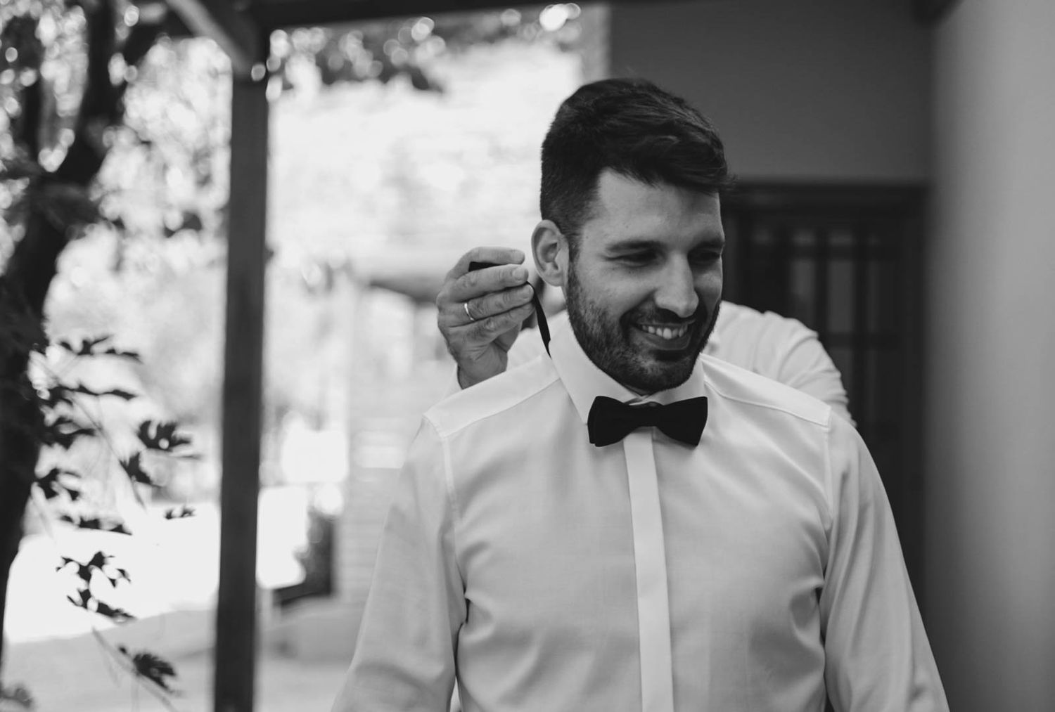 A black and white photo shows a person in formal attire with a bowtie smiling outdoors.