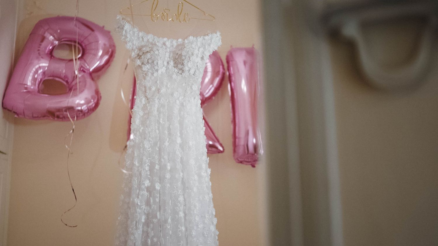 A delicate white lace wedding dress hangs on display with pink balloon letters visible in the background.