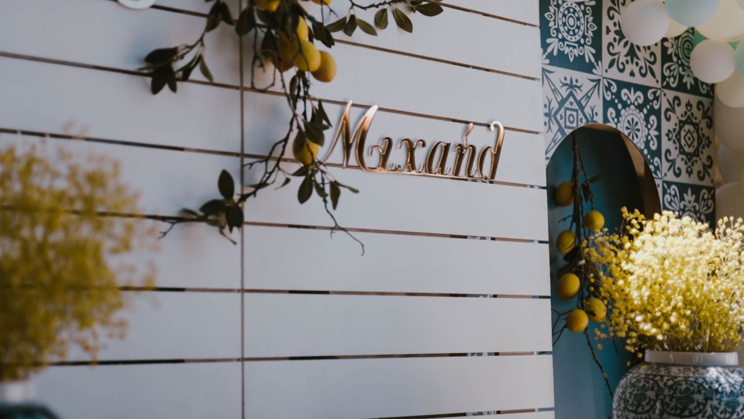 A white weatherboard wall with a decorative text sign, blue door frame, and fresh yellow flowers in a vase.