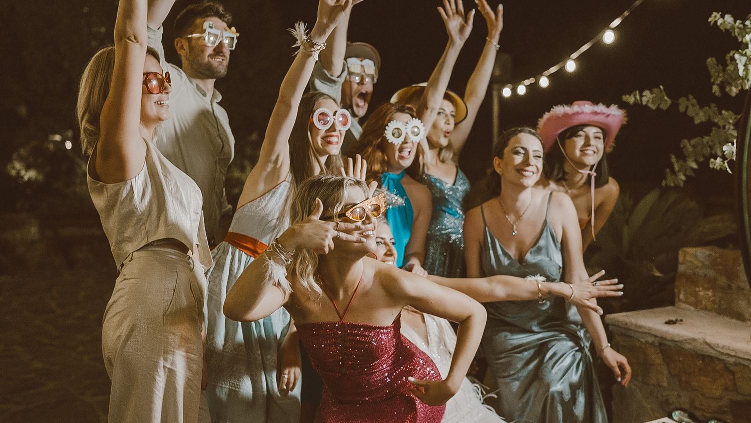 Group of friends having fun and dancing together at an outdoor party with string lights in the background.
