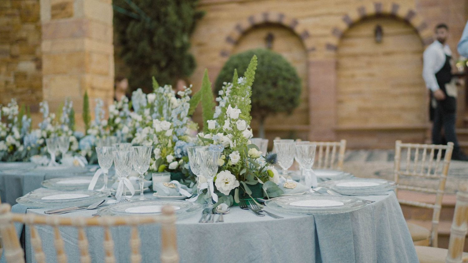 Elegant wedding table decor features white floral centerpieces and soft blue linens at an outdoor venue with stone archways.
