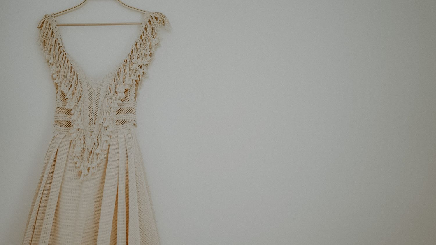 Elegant cream-colored wedding dress with lace details hanging against white wall.