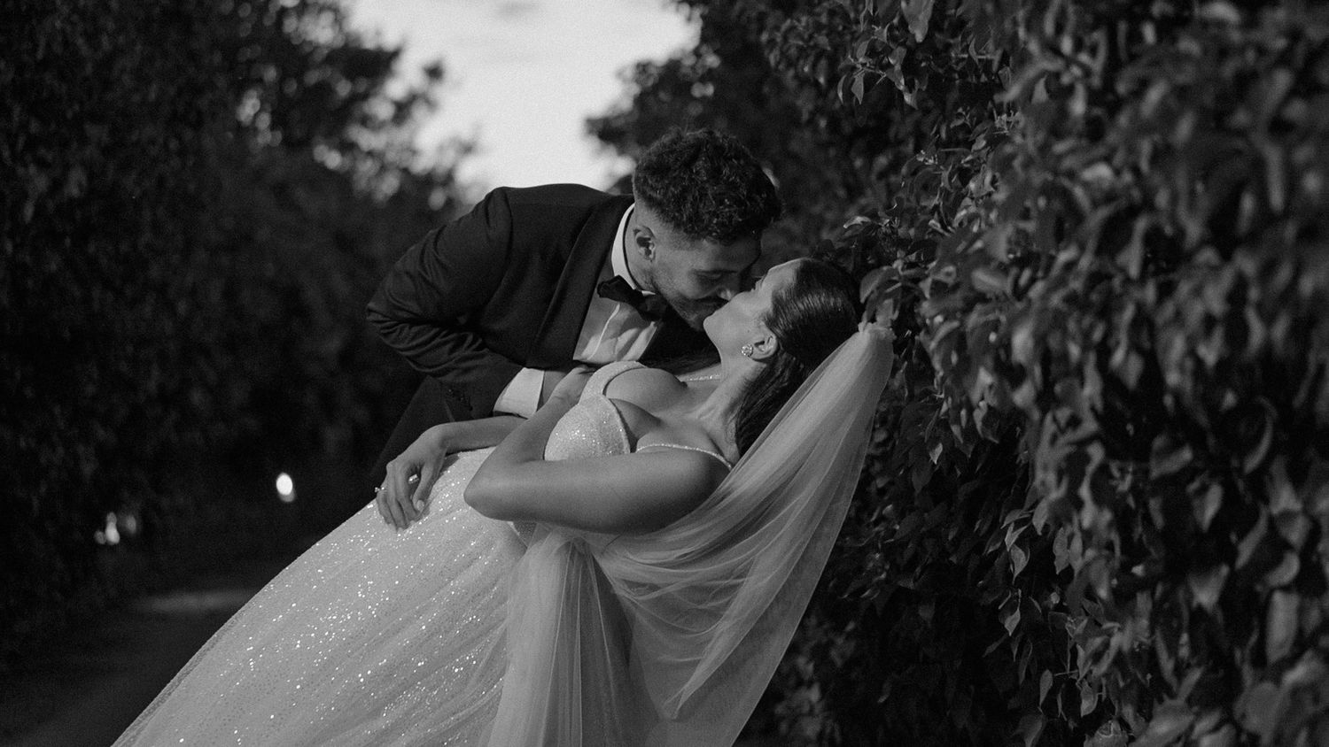 A romantic black and white wedding photo shows a couple in an intimate embrace among garden foliage.