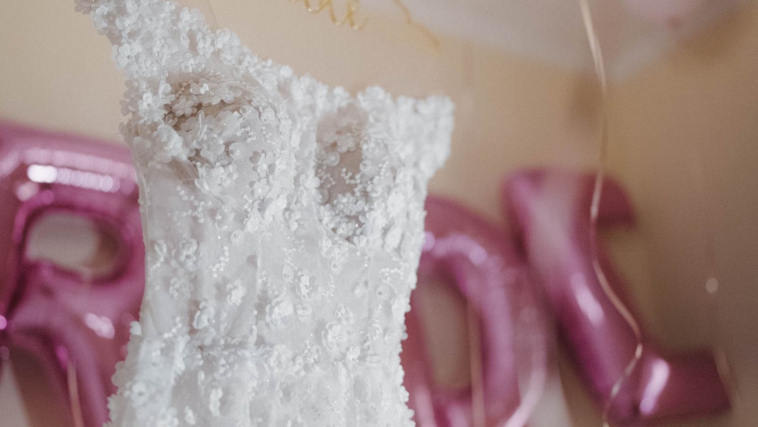 Close-up detail of a white lace wedding dress bodice with delicate floral patterns and pink ribbons in the background.