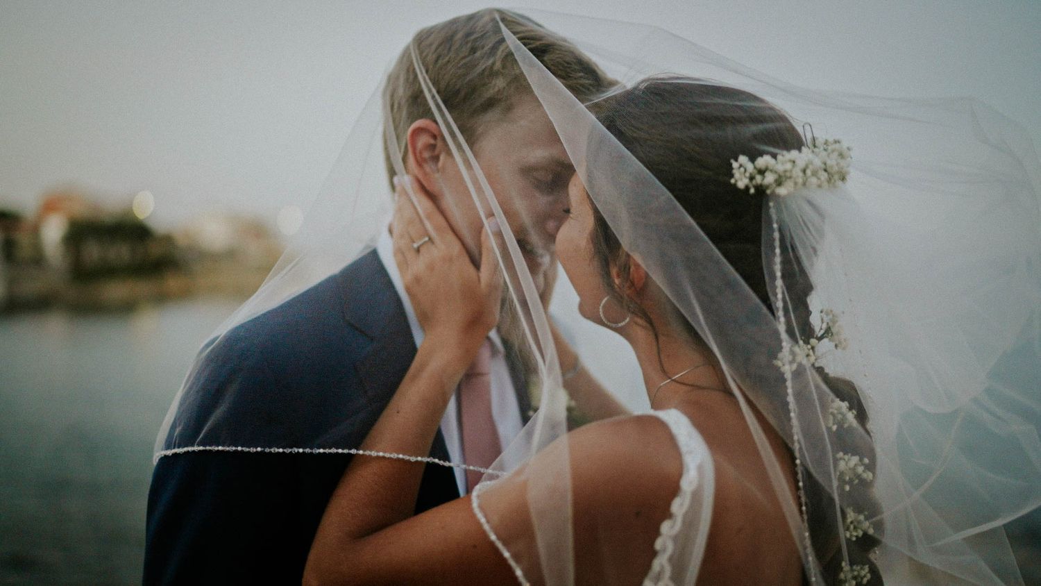 Romantic wedding couple embraces under flowing veil against waterfront backdrop at sunset.