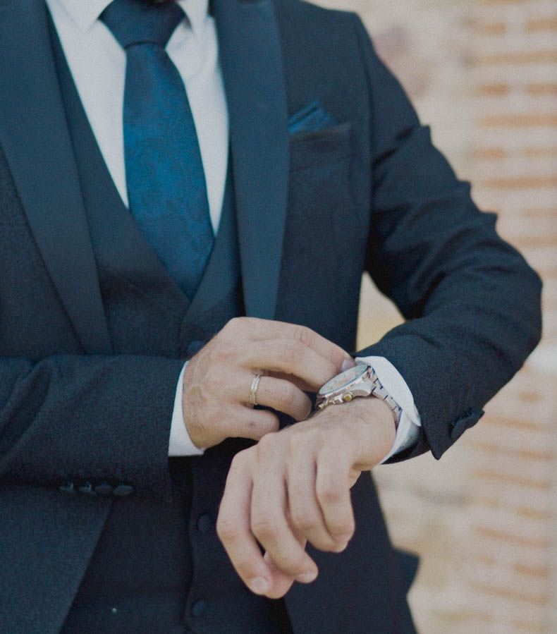 Close up of a person in navy suit and blue tie checking their wristwatch.