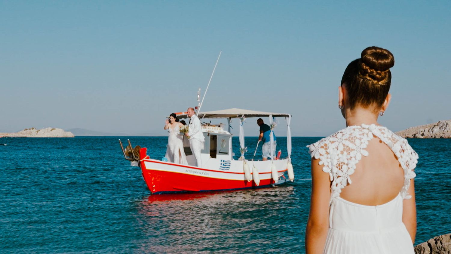 A person in a white lace dress watches a red and white fishing boat on bright blue Mediterranean waters.