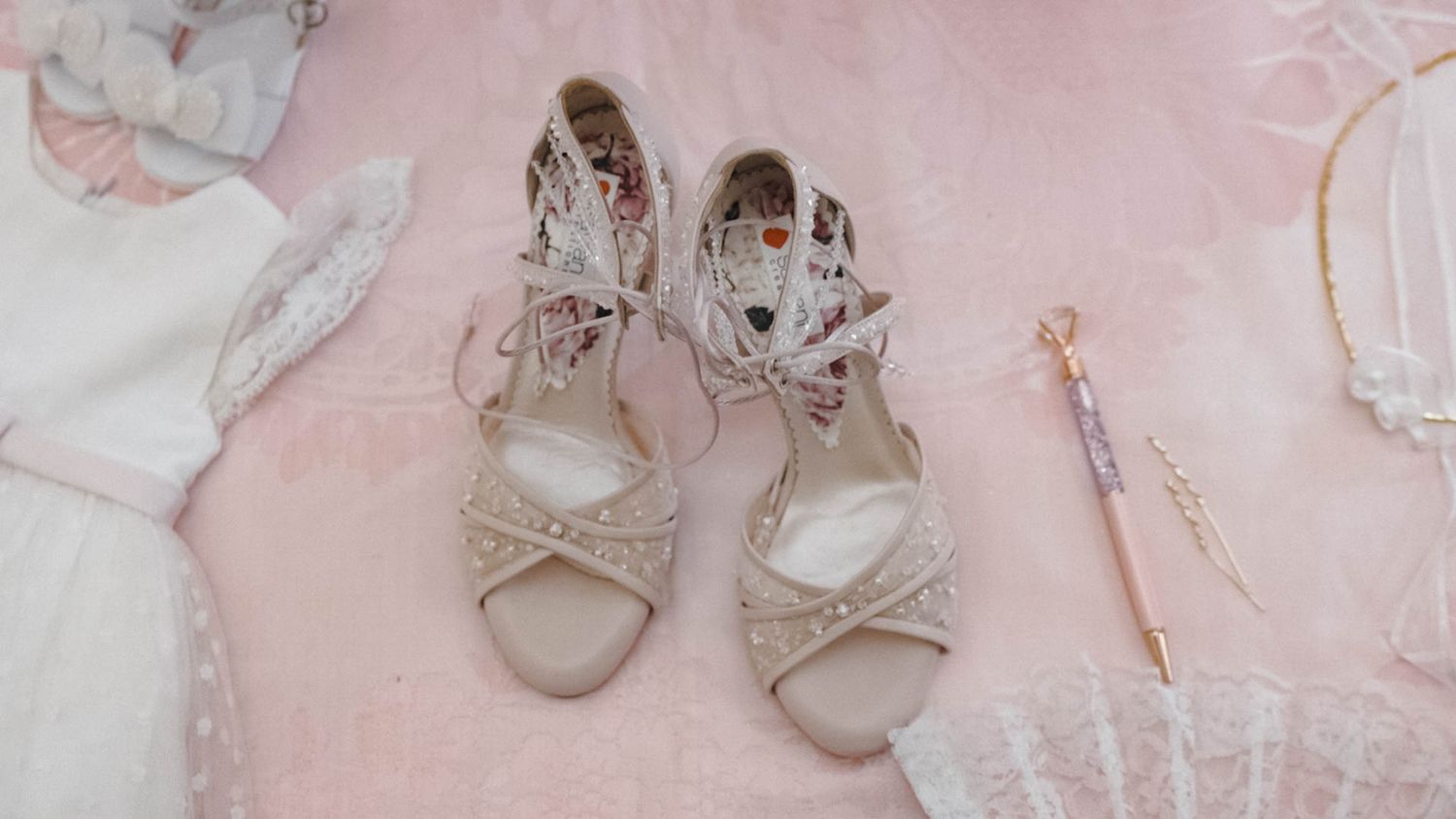 Elegant cream-colored bridal sandals with strappy design displayed on pink fabric with delicate lace accents.