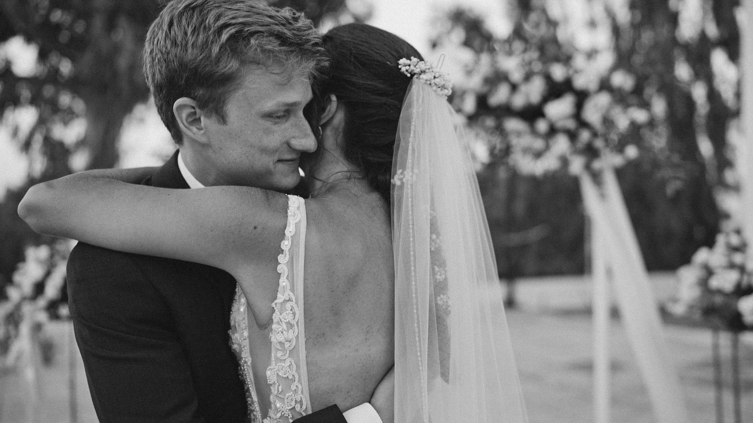A black and white photograph captures a newlywed couple embracing during their wedding ceremony in an outdoor garden setting.