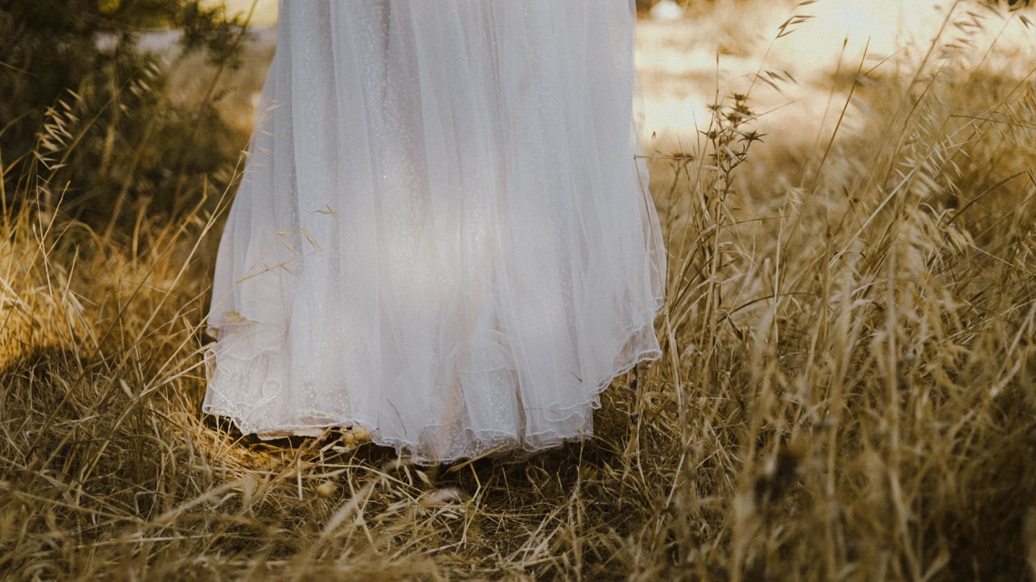 Bottom of a flowing white wedding dress visible among tall golden grass at sunset.