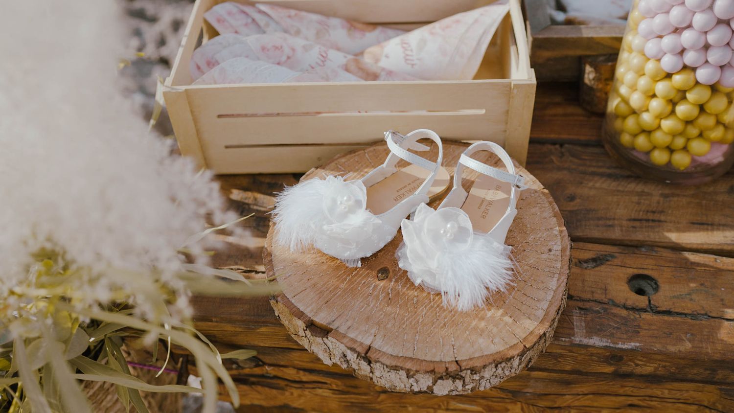 White knit baby booties with pom poms displayed on a rustic wood slice with floral decor.
