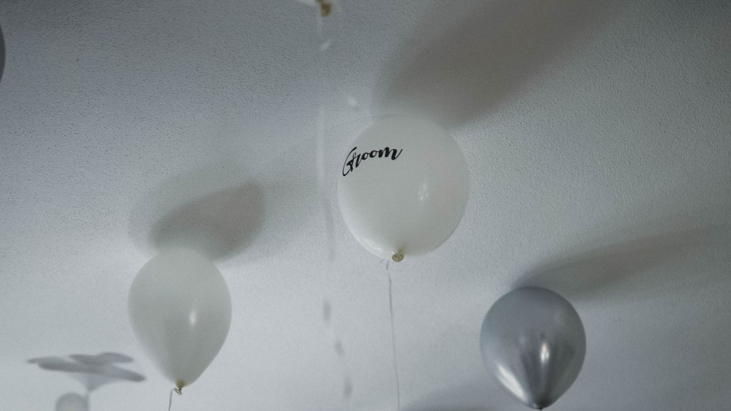 White and gray helium balloons floating against a soft white ceiling with gentle shadows cast above.