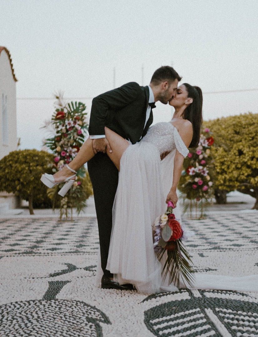 A couple shares a romantic kiss on a tiled patio with blooming flowers in the background.