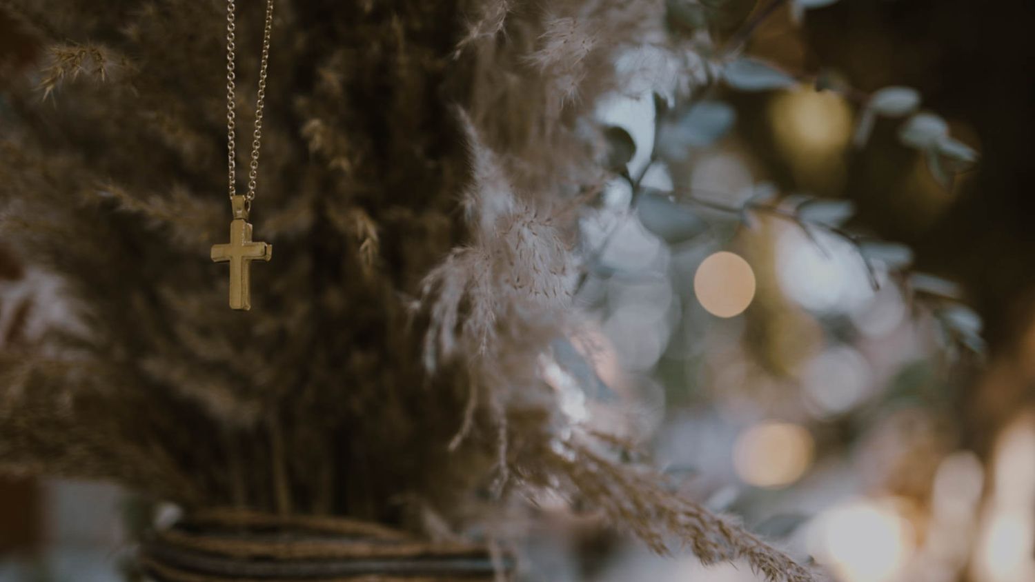 A gold cross necklace pendant hangs against a dark background with soft bokeh lights and smoke effects.