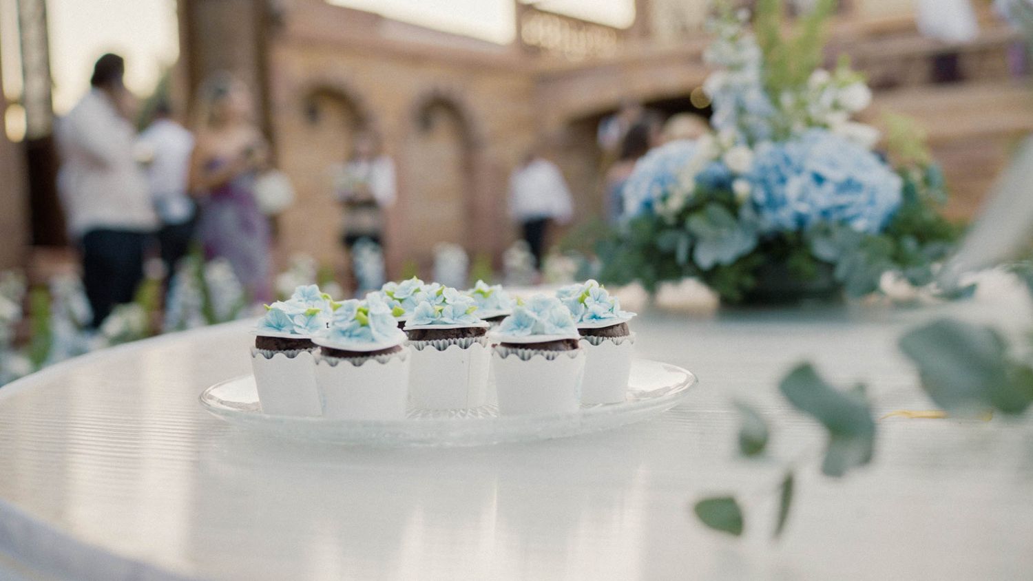 Fine china teacups with floral arrangements sit elegantly on a white tablecloth at an indoor event setting.