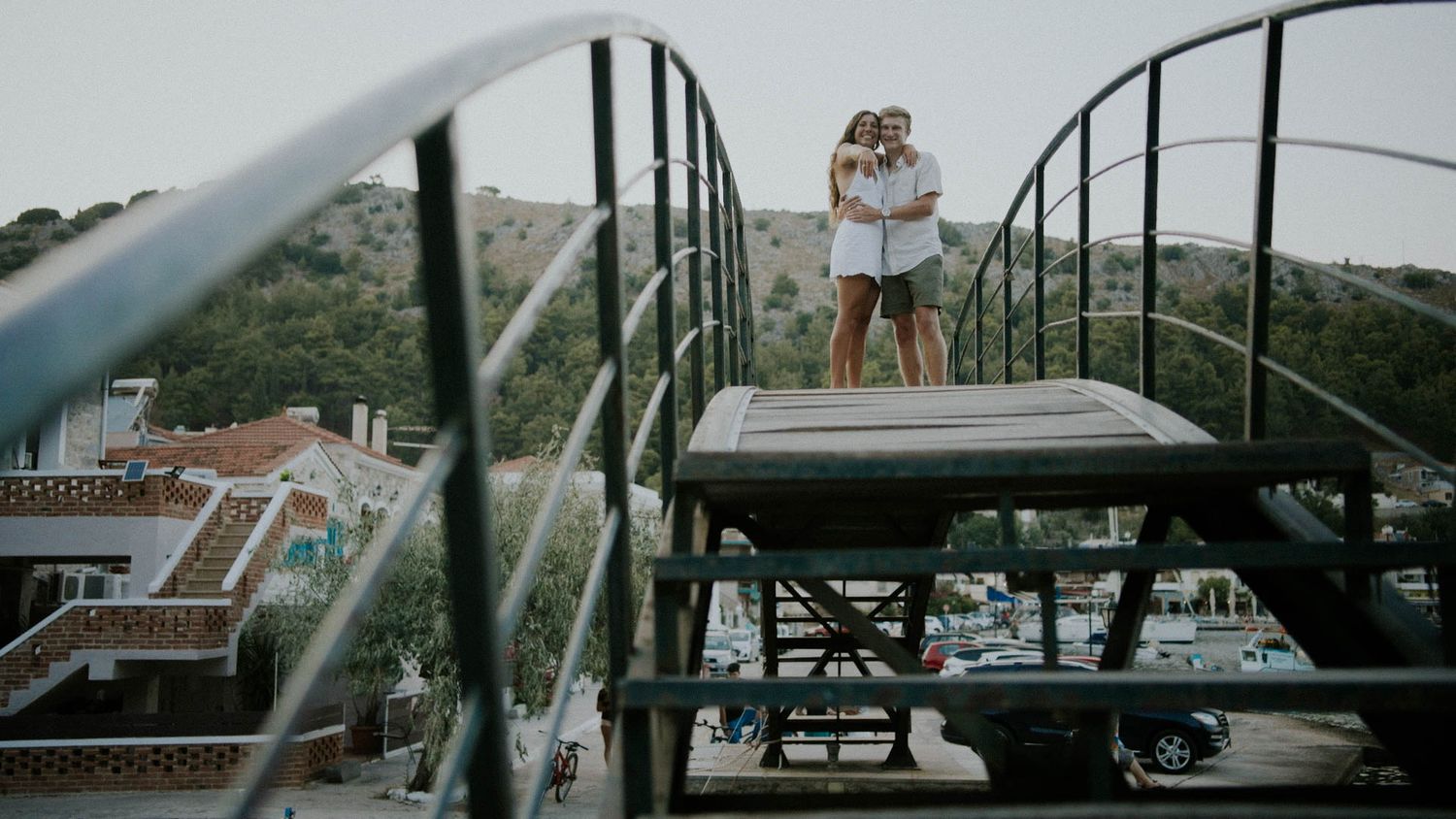 Two people embrace on a steel railway bridge overlooking a scenic landscape with buildings and trees below.